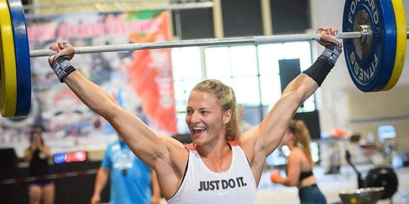 Female athlete performing an overhead lift with a barbell and wrist wraps in a gym.