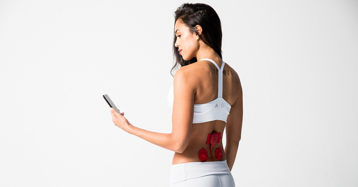 Woman in sportswear using red muscle stimulator pads on lower back while holding phone