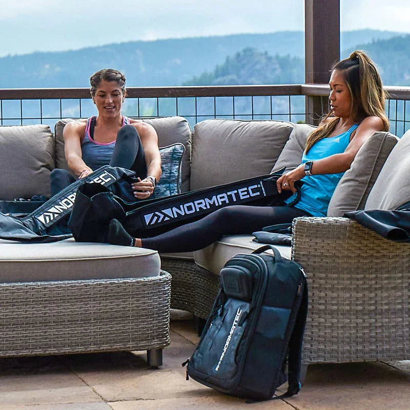 Two women using Normatec compression boots for recovery on an outdoor patio sofa.