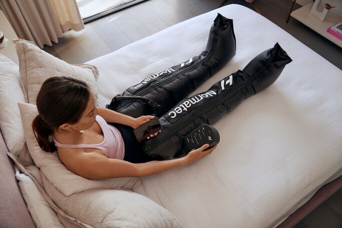 Woman using Normatec leg recovery compression sleeves on bed for athletic muscle recovery
