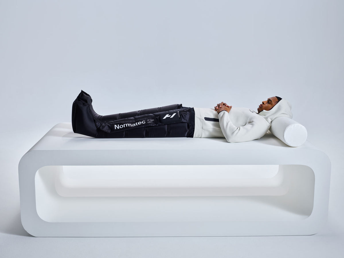 A person wearing Normatec compression boots and a white hoodie lying on a modern white bench.