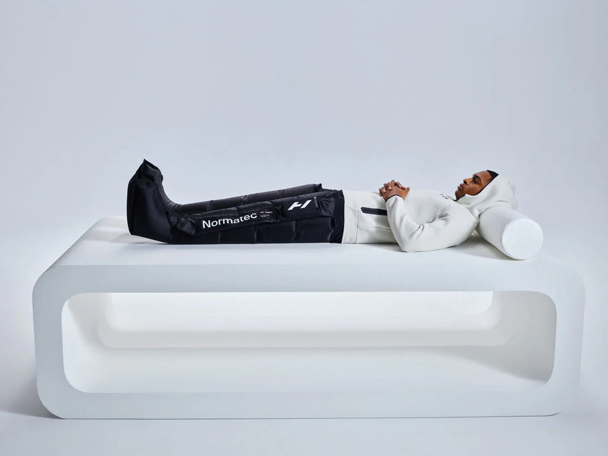 Person lying on a white platform wearing Normatec compression recovery boots and a white hoodie.