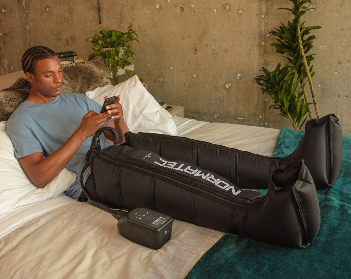 A man relaxing on a bed using Normatec compression leg recovery boots while on his phone.