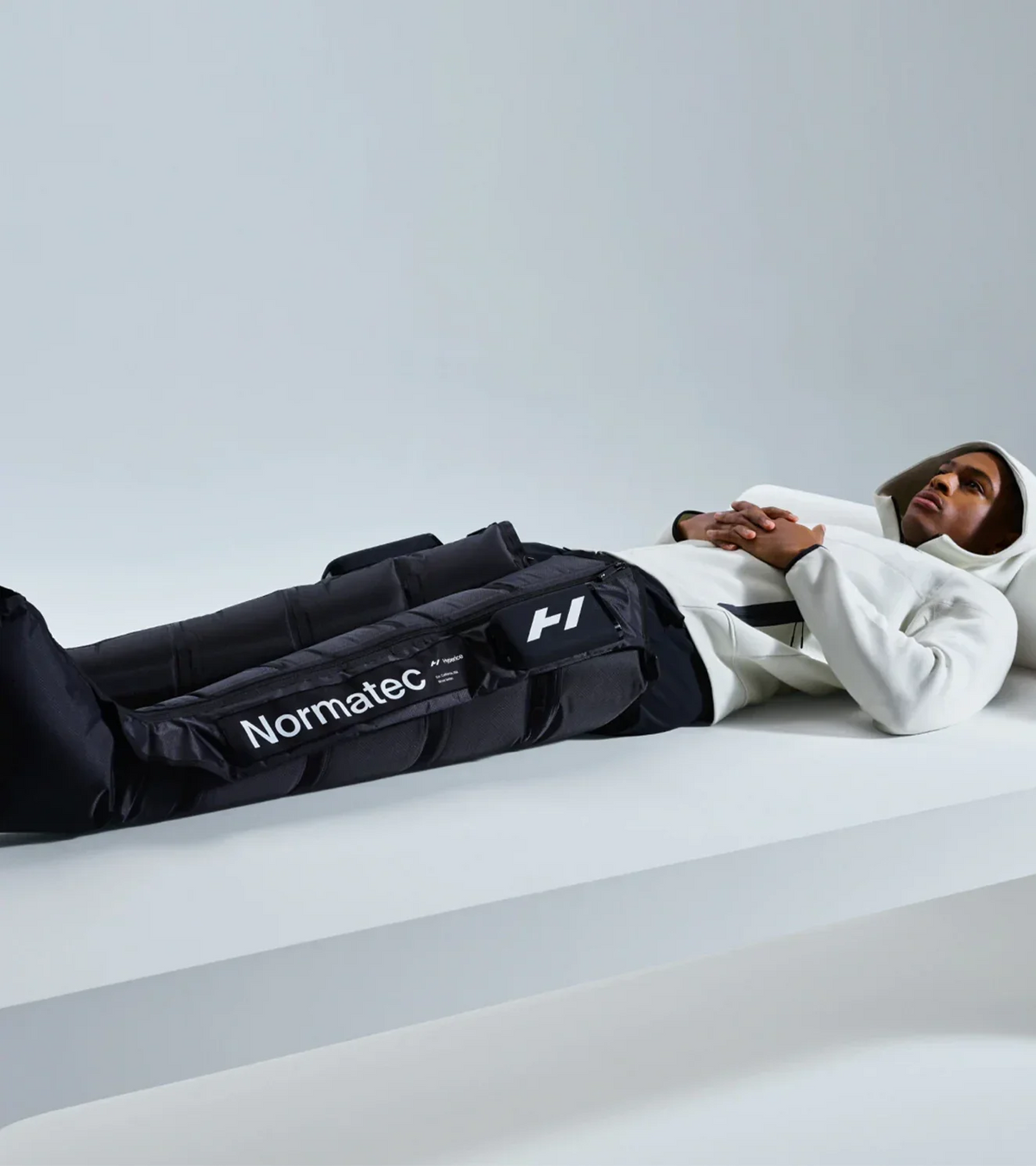 Person lying down wearing black Normatec recovery compression boots and a white hoodie.