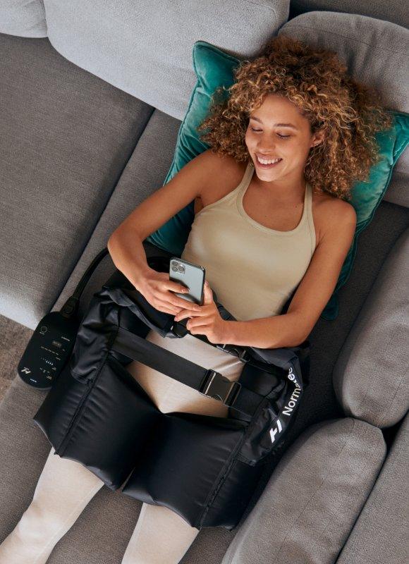 Woman relaxing on sofa using leg recovery compression boots, holding a smartphone