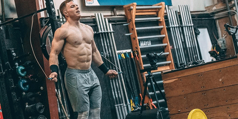 Athletic man jumping rope shirtless in a functional fitness gym with equipment in the background