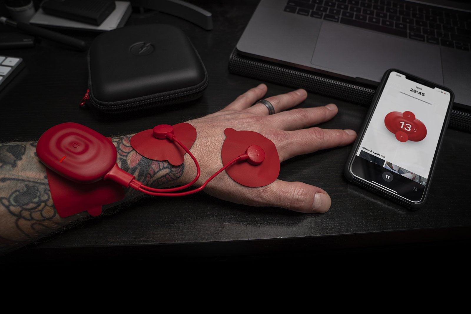 Red muscle stimulator device on tattooed forearm, connected to mobile app, on desk with laptop.