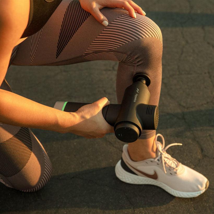 Athlete using Hypervolt massage gun on leg outdoors, wearing patterned leggings and sneakers