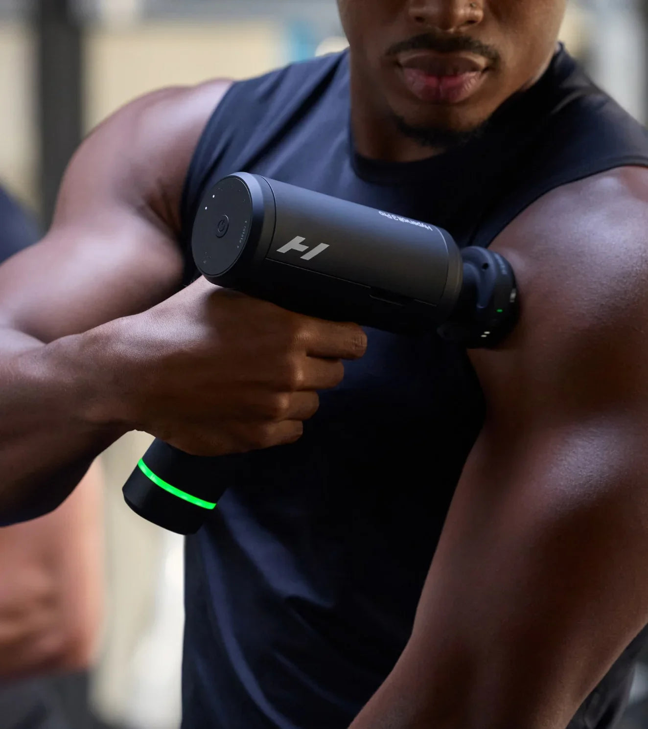 Athlete using black massage gun on shoulder, green LED light, fitness recovery tool for muscle relief