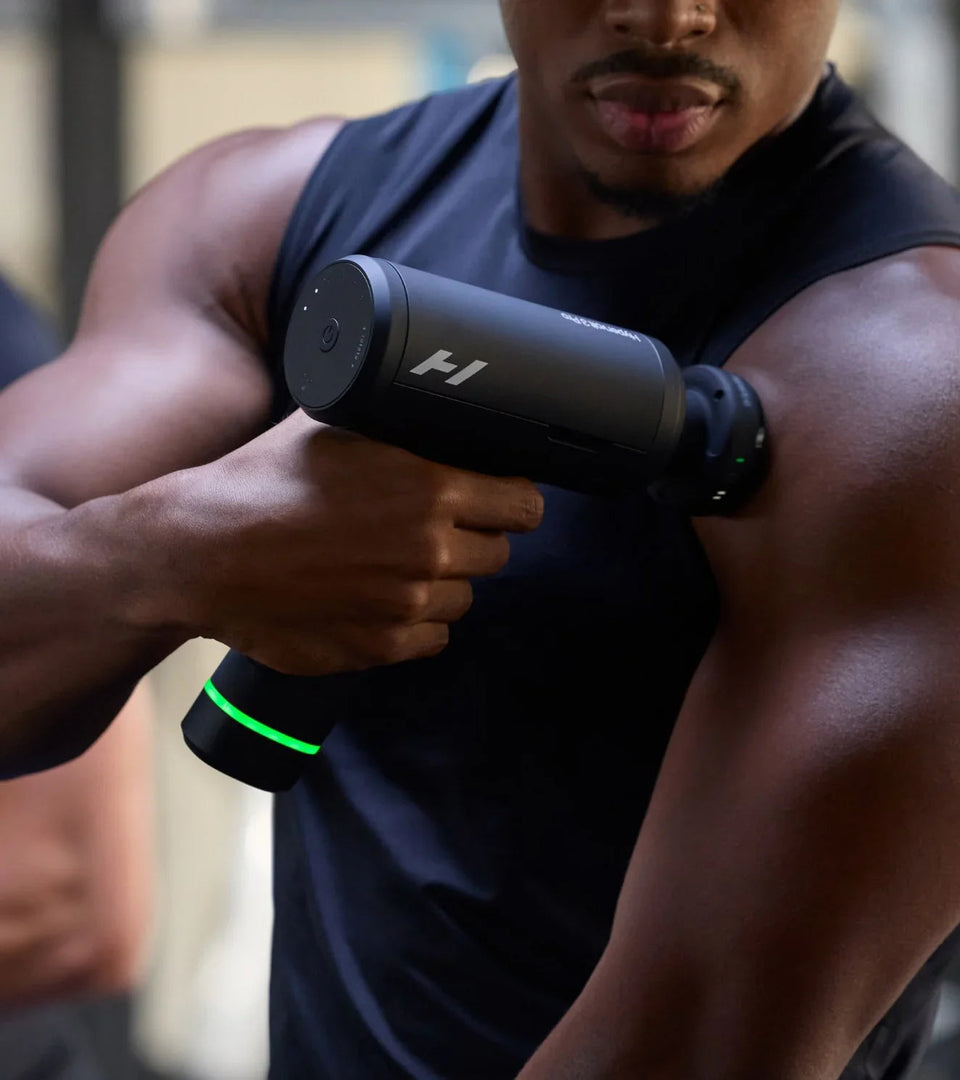 Athlete using black massage gun on shoulder, green LED light, fitness recovery tool for muscle relief