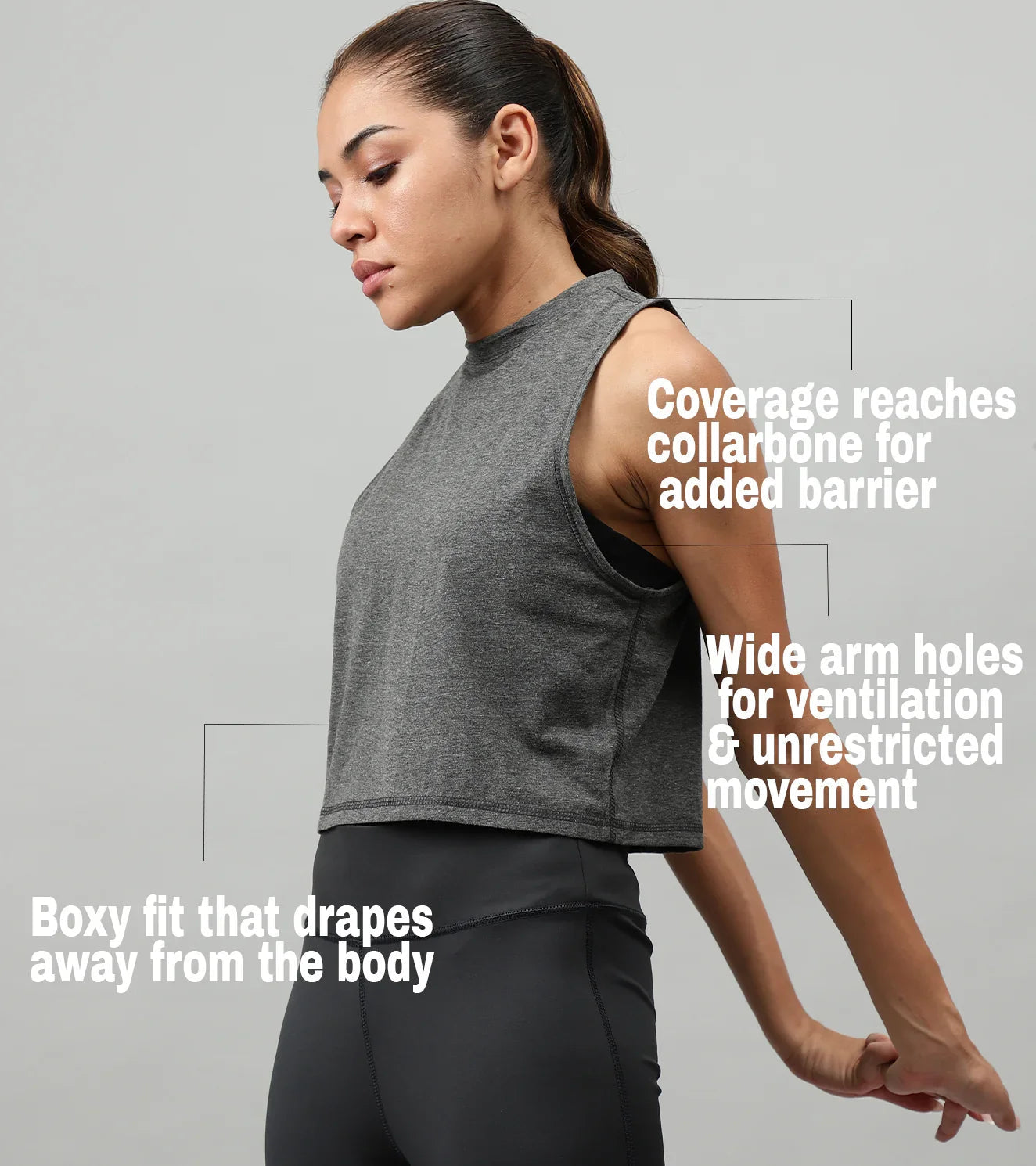 Woman wearing a gray sleeveless athletic crop top with coverage to collarbone, boxy fit, and wide arm holes, stretching arms in a fitness studio.