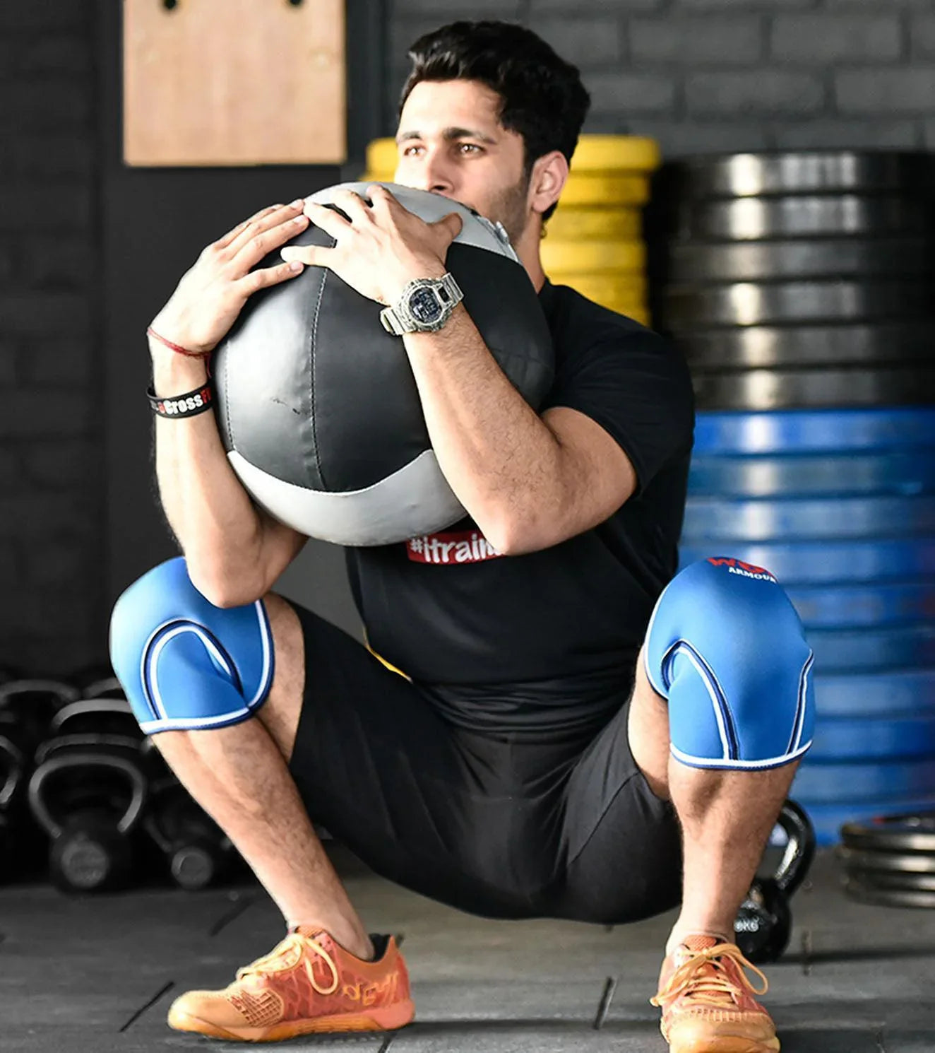 Man in a gym wearing blue knee sleeves holding a weighted medicine ball in a squat position.