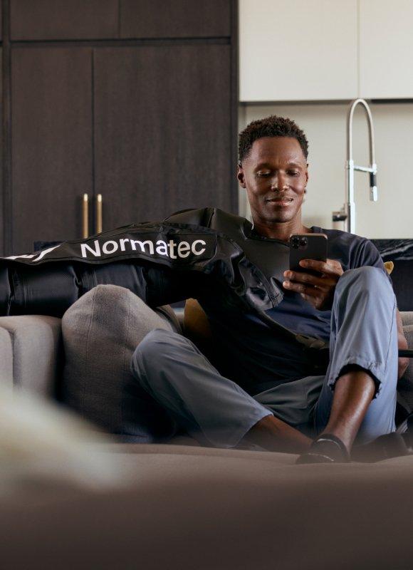 Man using Normatec compression recovery boots on couch, relaxing with smartphone indoors