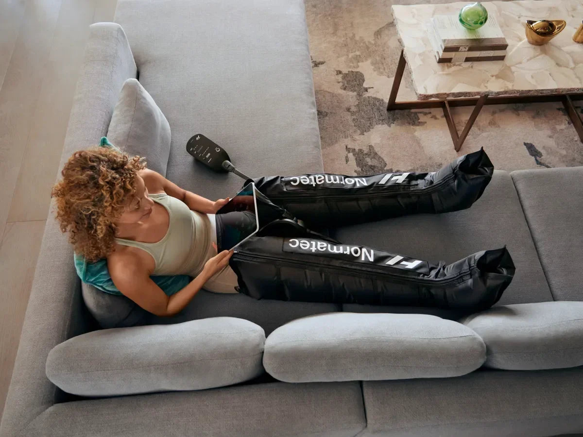 Woman relaxing on a couch using Normatec compression leg boots for recovery.