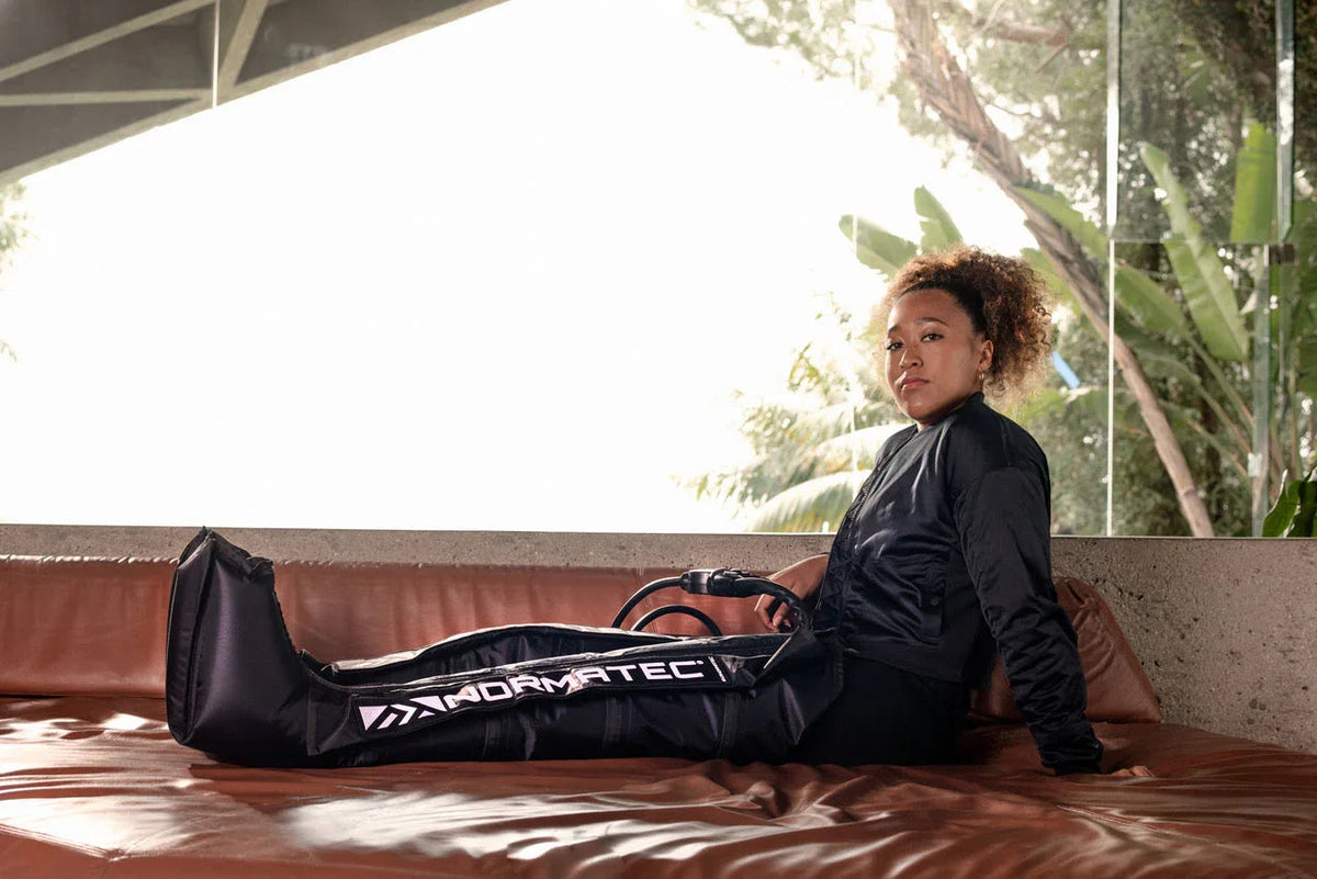 Woman resting on a leather sofa using Normatec compression boots for athletic recovery.