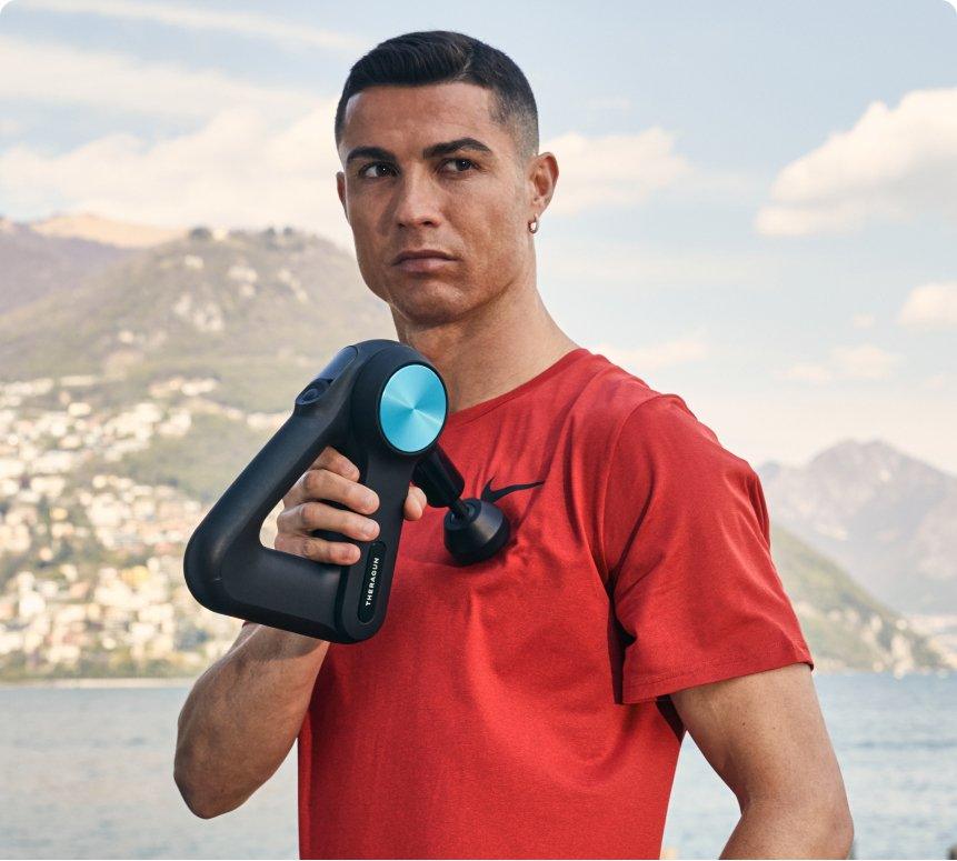 Athlete using a handheld massage gun outdoors with mountains and water in background