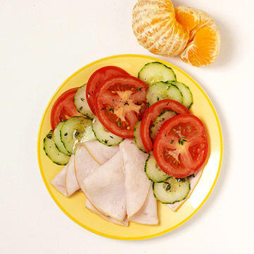 Healthy plate of sliced turkey, tomatoes, cucumbers, and a peeled orange for a fitness diet