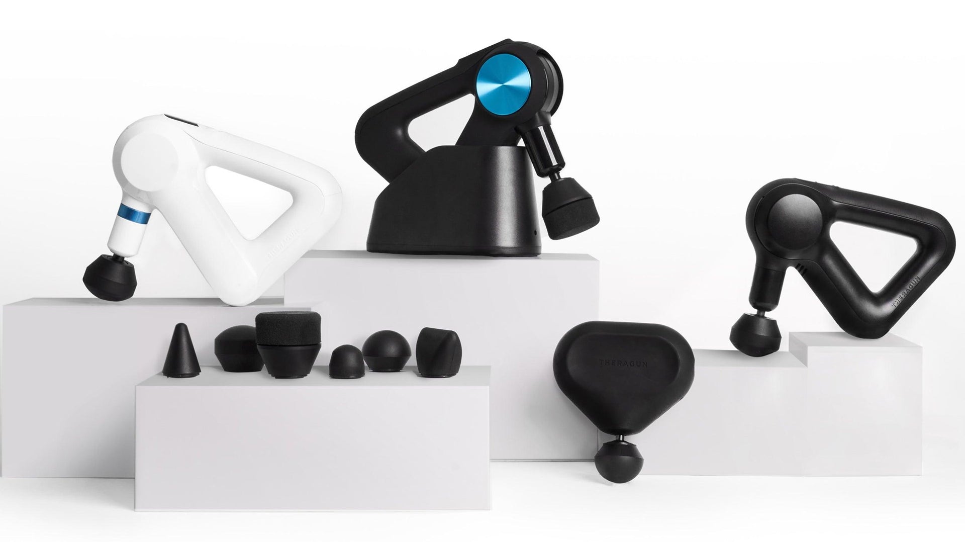 Four Theragun massage guns with different attachments on white display blocks for muscle recovery