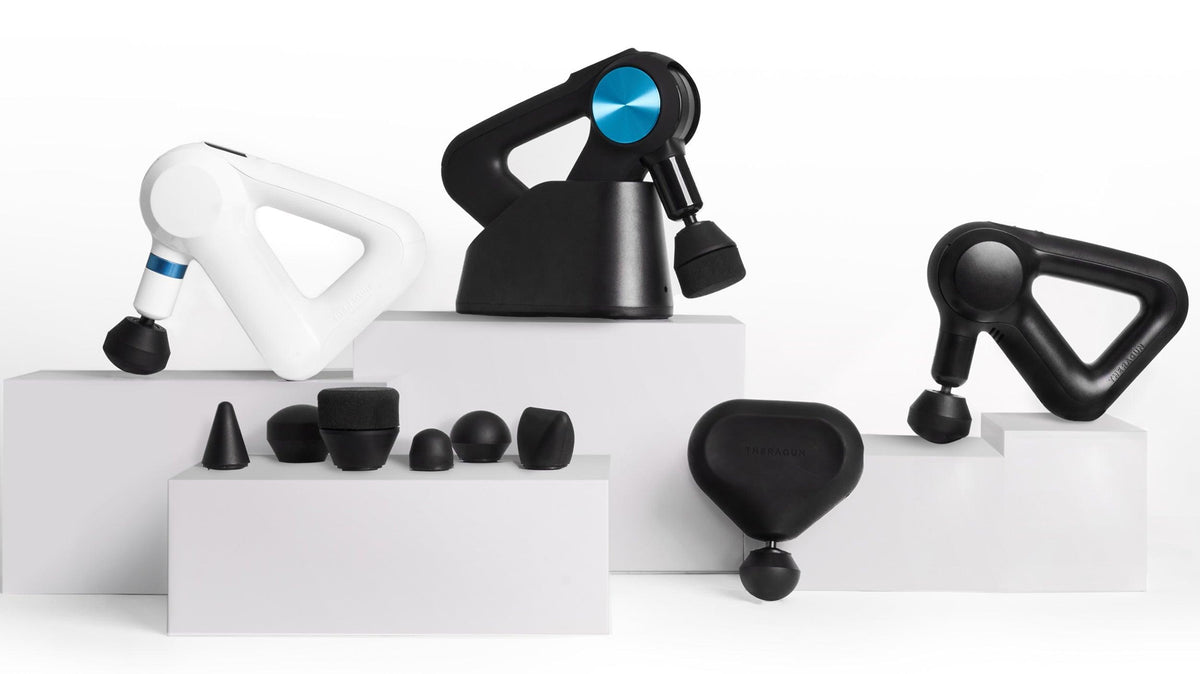 Collection of Theragun massage guns and interchangeable attachments on white pedestals.