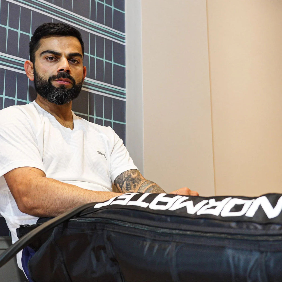 Virat Kohli in a white t-shirt sitting next to a Normatec recovery boot bag.