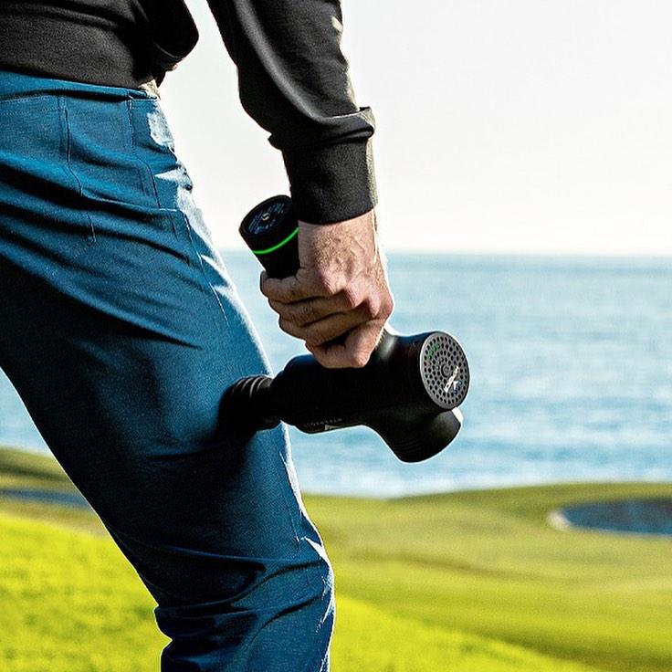 Athlete using a percussive massage gun on thigh outdoors by the water