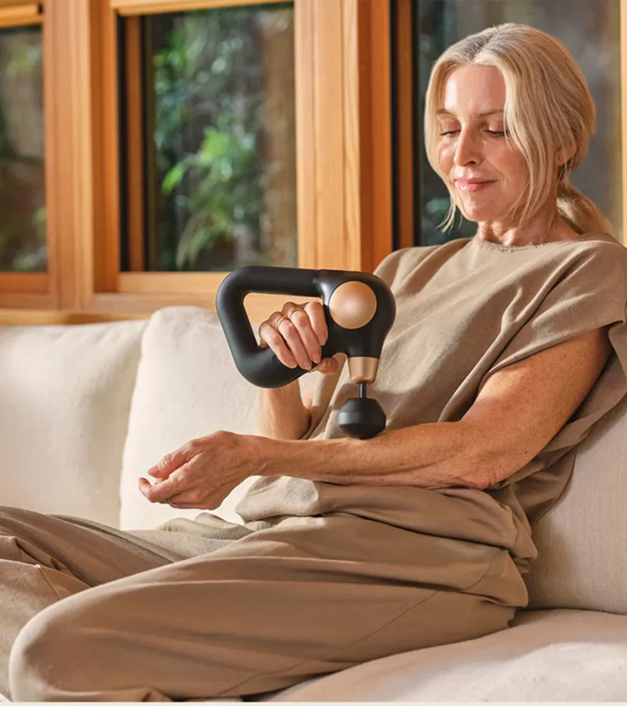 Woman using handheld massage gun on arm for muscle recovery at home