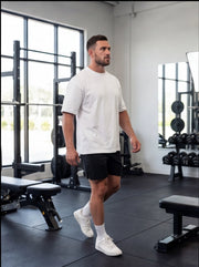 Man in white tee and black shorts walking in gym with weight racks and benches