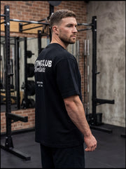 Man in black gym t-shirt with 'Lifting Club Worldwide' print, standing in functional fitness gym with squat racks