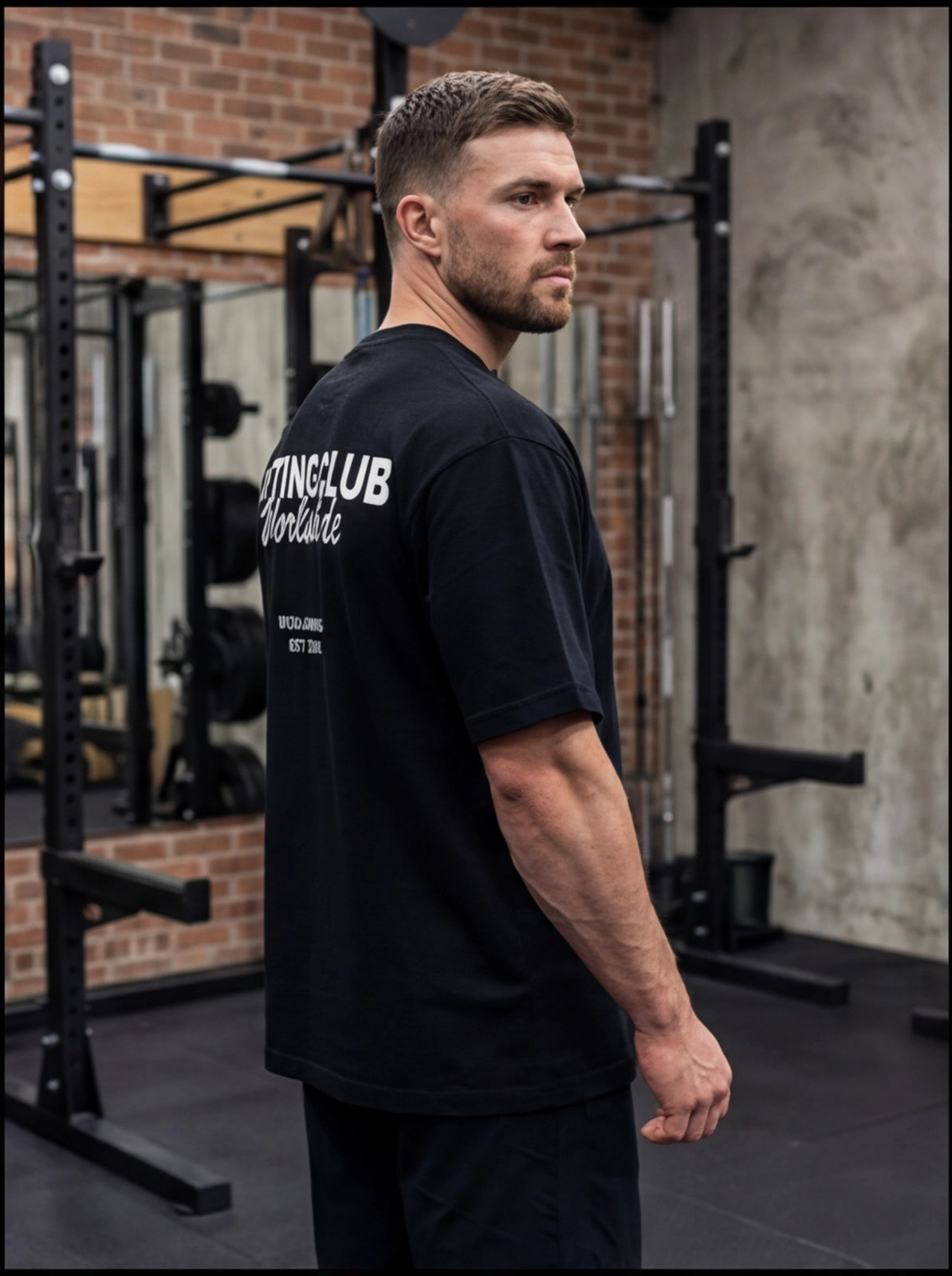 Man in black gym t-shirt with 'Lifting Club Worldwide' print, standing in functional fitness gym with squat racks