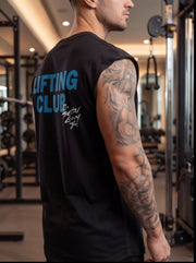 Man in gym wearing black sleeveless 'LIFTING CLUB' shirt, showcasing tattooed arm during workout.
