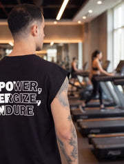 Man in black sleeveless gym shirt with 'POWER, ENERGIZE, ENDURE' text, tattooed arm, in fitness studio with treadmills