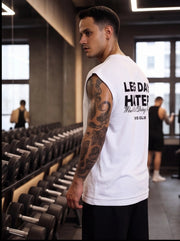 Man in white 'Leg Day Hater' gym tank top, tattooed arm, standing by dumbbell rack in fitness studio