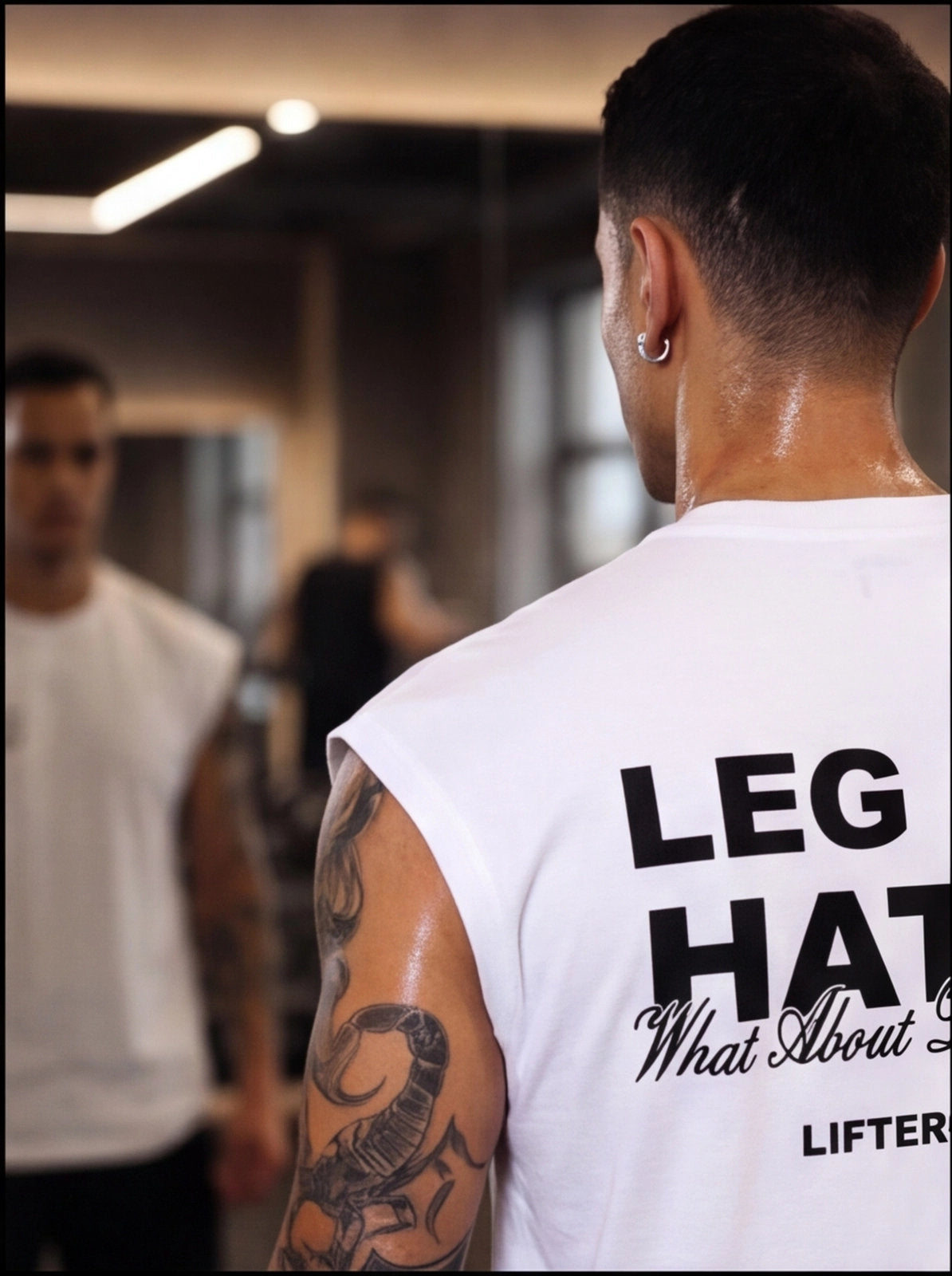 Man in gym wearing white 'LEG HAT' tank top, tattooed arm, sweat after workout