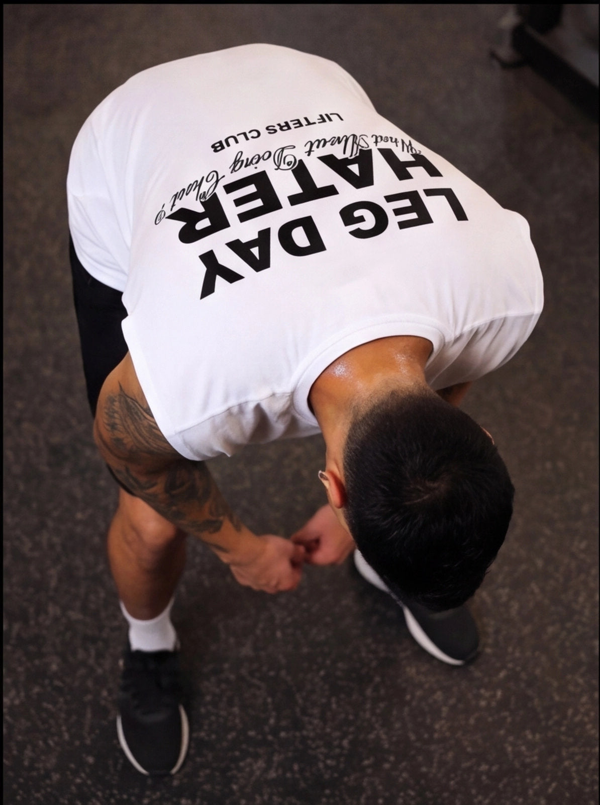 Man in white 'Leg Day Hater' gym shirt, bending over, tattooed arm, workout gear