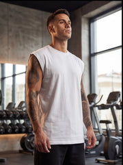 Man in white sleeveless gym shirt, tattooed arms, standing in fitness studio with weights and cardio machines