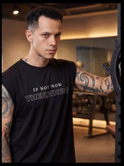 Man in gym wearing black sleeveless fitness tee with 'IF NOT NOW THEN, WHEN?' slogan, motivational workout apparel