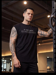 Man in gym wearing black sleeveless fitness shirt with 'IF NOT NOW THEN WHEN?' text, tattoos visible, leaning on weight rack.