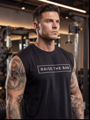 Man in gym wearing black 'Raise The Bar' sleeveless fitness shirt, showcasing athletic build and tattoos.