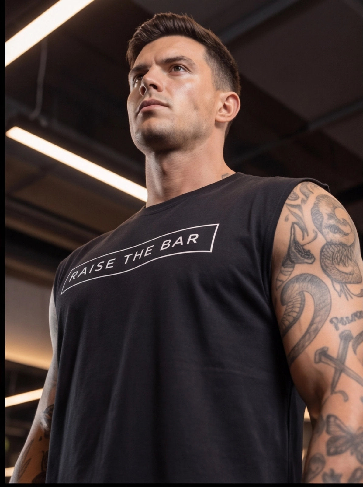 Man in gym wearing black 'Raise The Bar' sleeveless fitness shirt, tattooed arms, motivational workout apparel