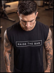 Man in gym wearing black 'Raise The Bar' sleeveless fitness shirt, showcasing athletic tattooed arms.
