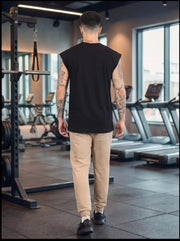 Man in black sleeveless gym shirt and tan joggers walking through fitness center with treadmills and squat rack