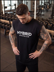 Man in black HYBRID ATHLETES CLUB sleeveless gym shirt, tattooed arms, hands on hips in fitness studio