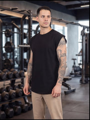 Man in black sleeveless gym shirt with tattoos, standing in fitness studio with dumbbells and equipment