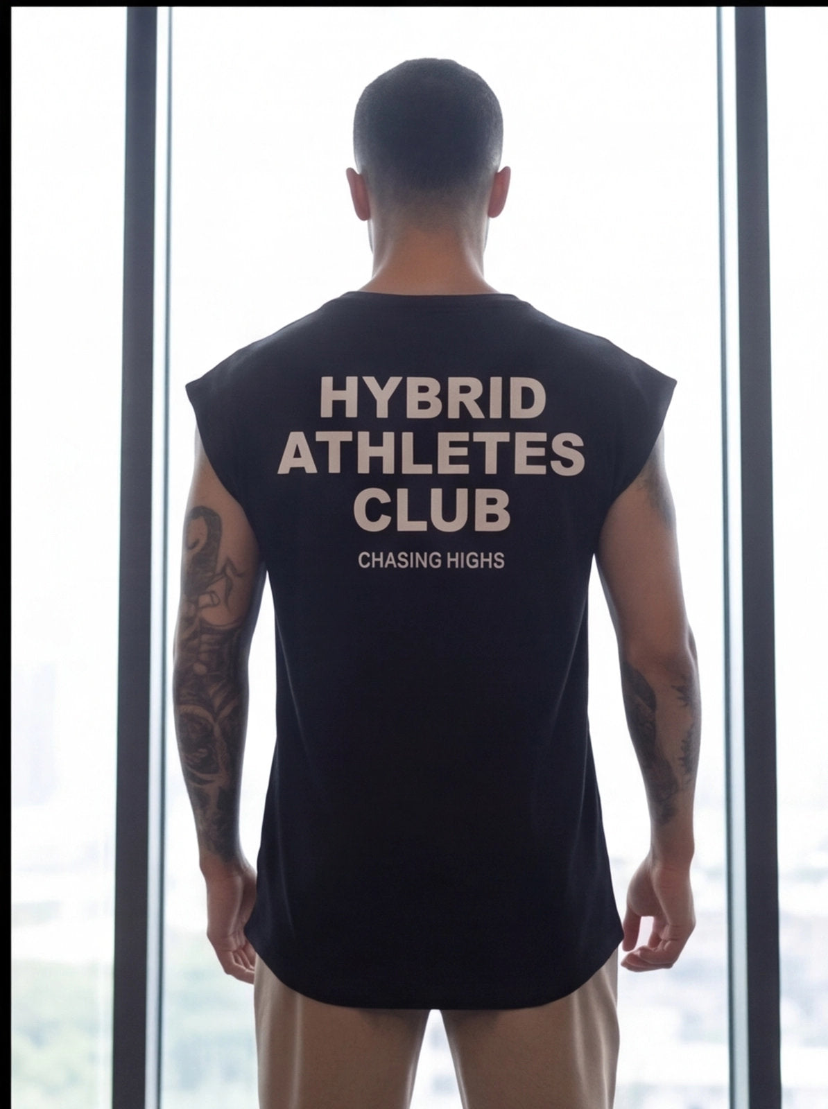 Man wearing black Hybrid Athletes Club sleeveless shirt, standing by window, fitness apparel for athletes.