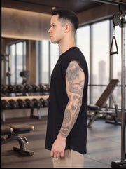 Man in black sleeveless gym shirt with tattooed arm, standing in fitness studio with dumbbells and equipment
