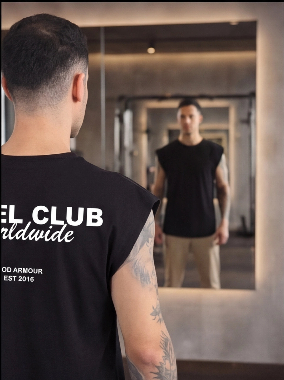Man in black sleeveless gym shirt with 'Fuel Club Worldwide' print, facing mirror in fitness studio