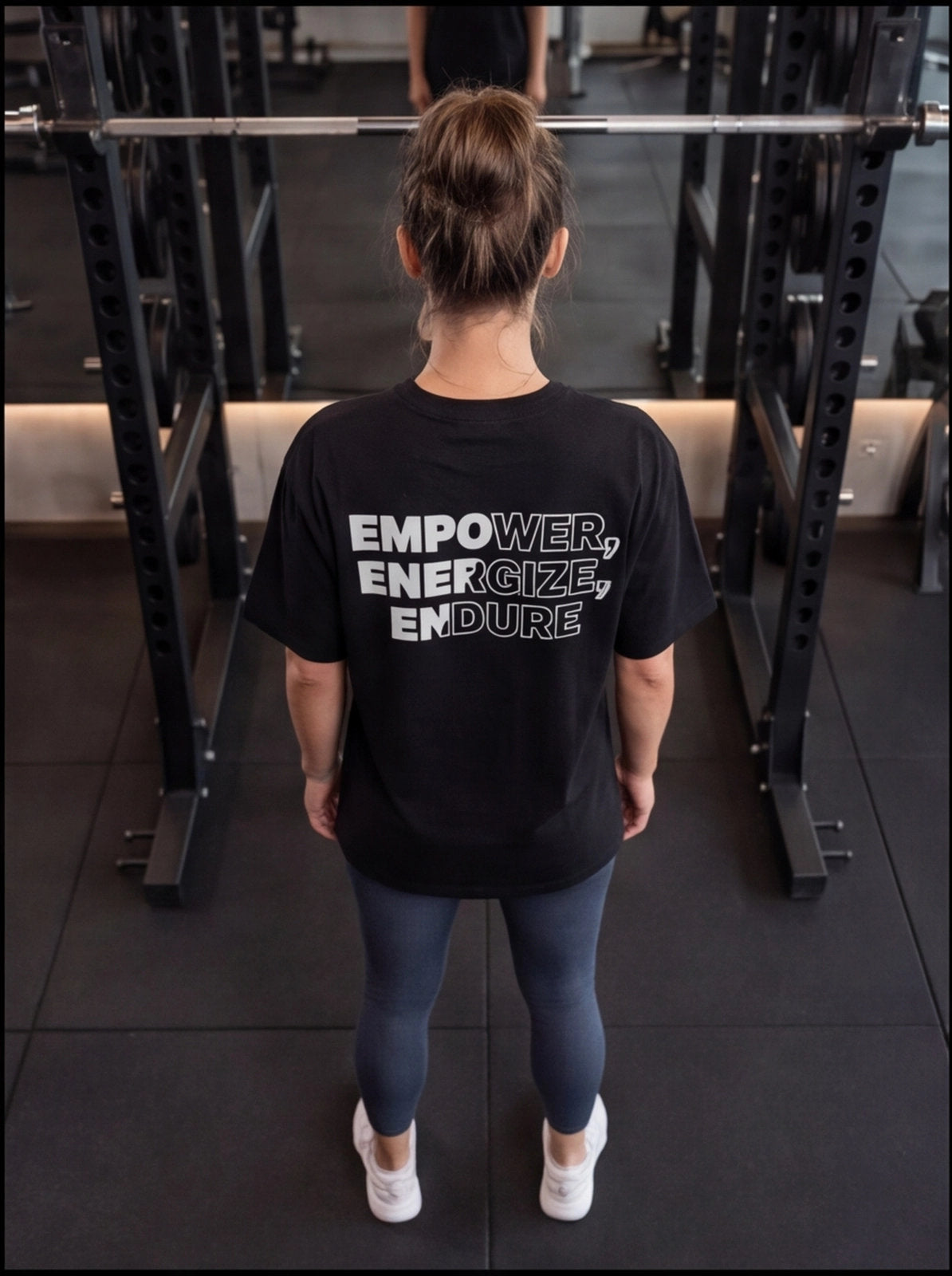 Woman in gym wearing black 'EMPOWER, ENERGIZE, ENDURE' t-shirt, leggings, white sneakers near squat rack