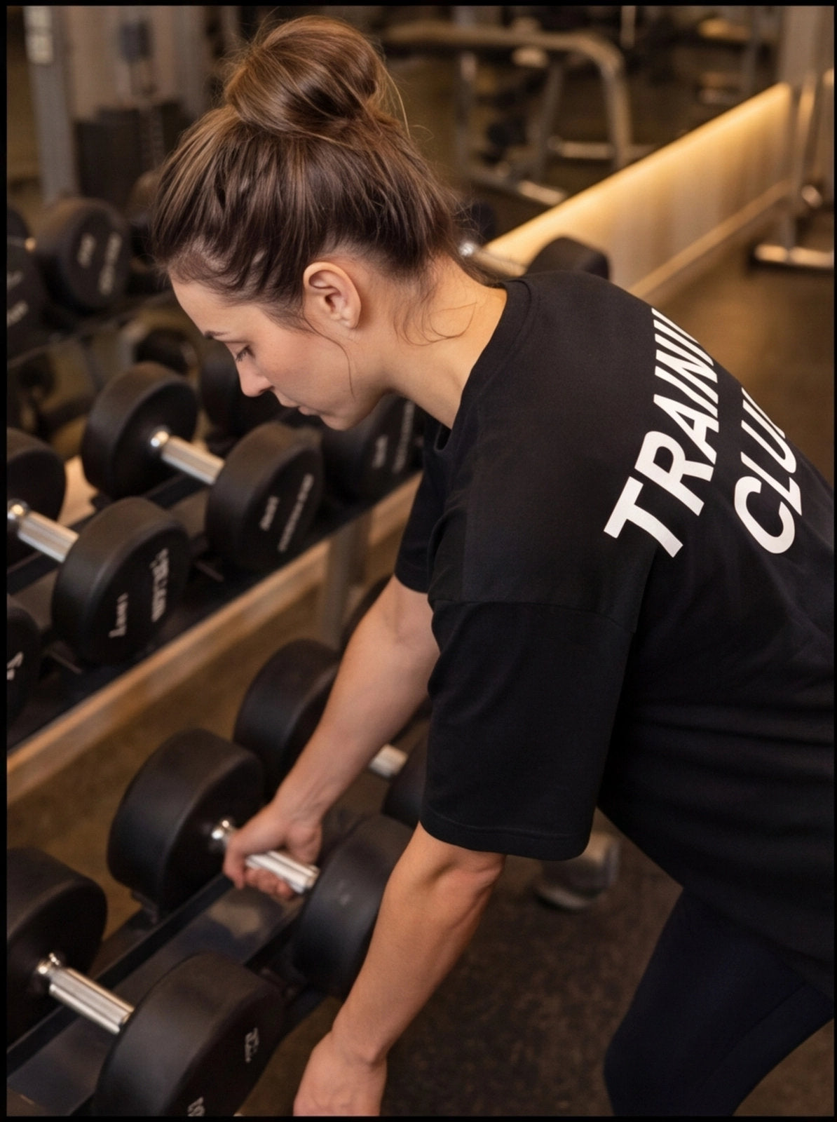 Woman in gym picking up dumbbell, wearing 'TRAINING CLUB' shirt for functional fitness workout