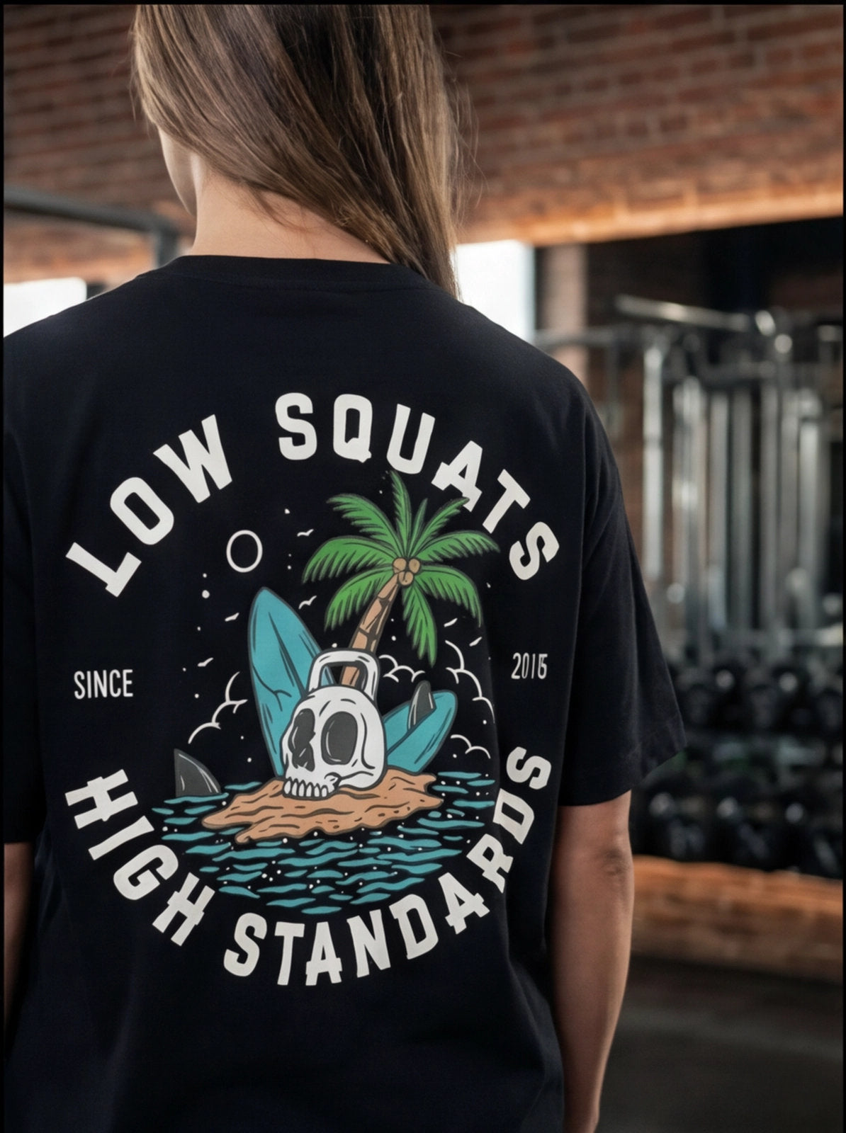 Black fitness tee with 'Low Squats High Standards' graphic, skull, palm tree, and surfboard, worn in gym.