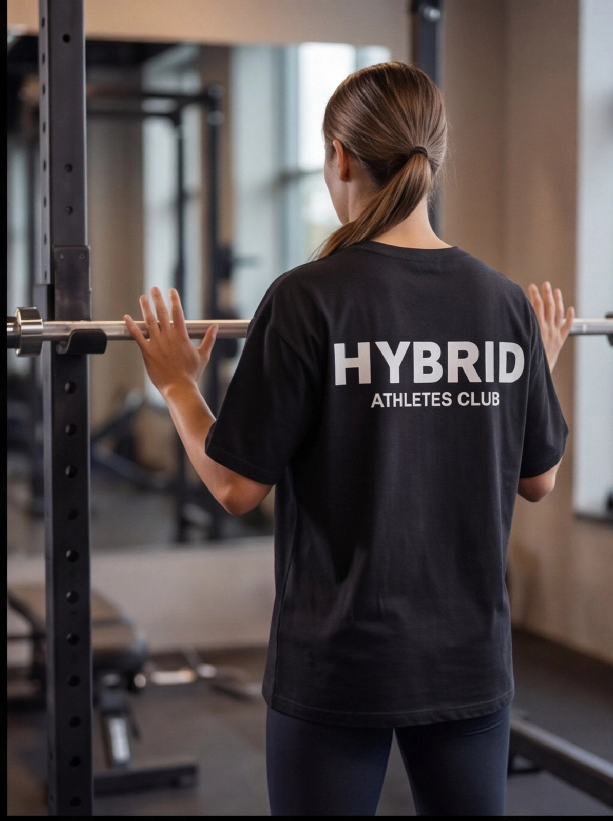 Woman in HYBRID ATHLETES CLUB tee at gym, hands on barbell, functional fitness apparel